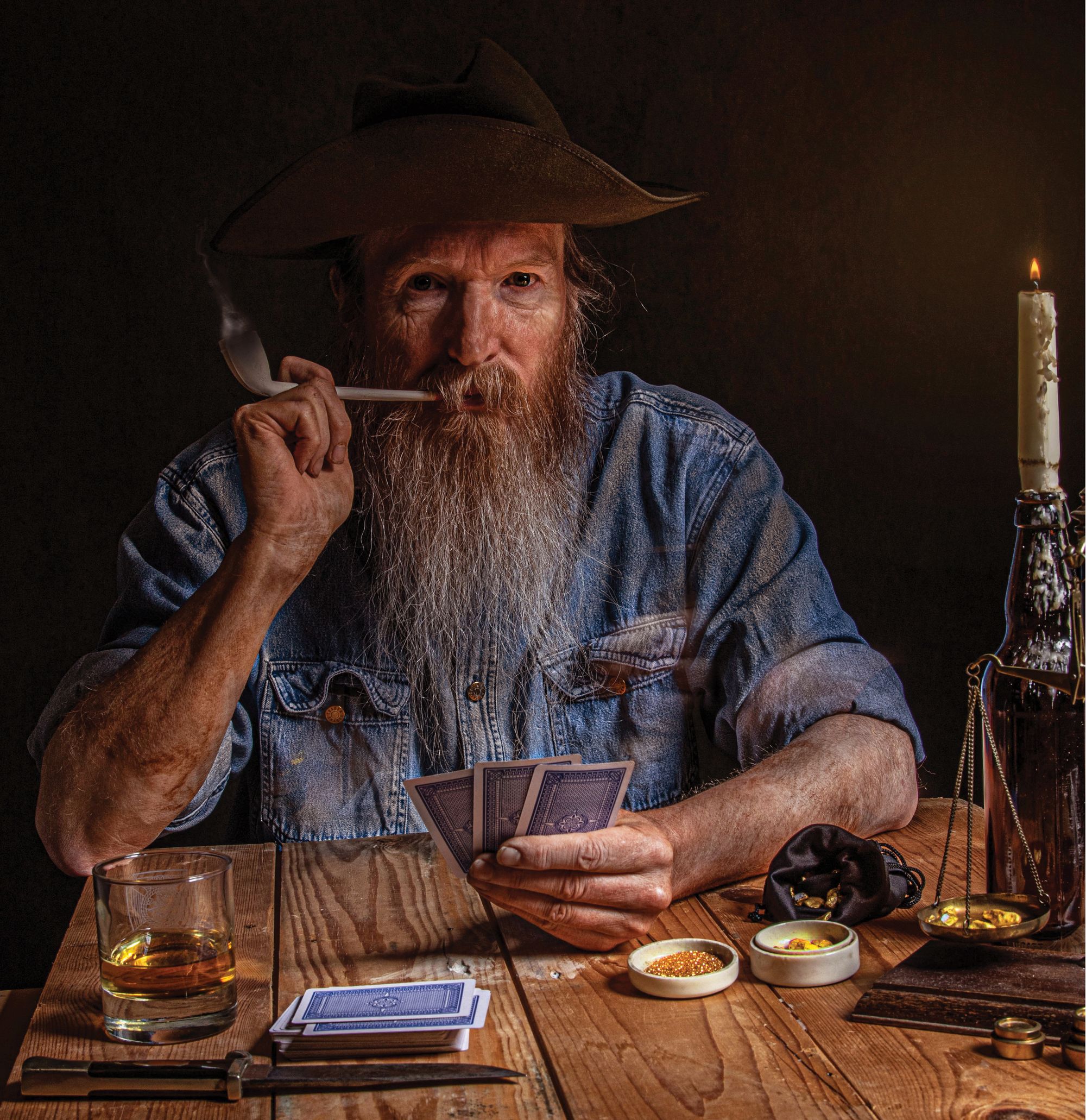 An old, bearded man in a blue denim shirt playing cards at a wooden table